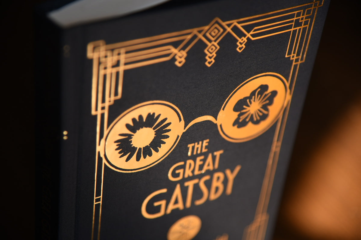 A close-up of a book cover for 'The Great Gatsby' featuring a black background with ornate gold Art Deco line art and typography.