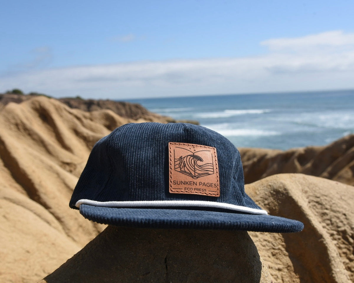 A navy blue corduroy snapback cap with a brown leather patch on the front that says Sunken Pages Eco Press, displayed outdoors on a rock with a sandy beach and ocean in the background.