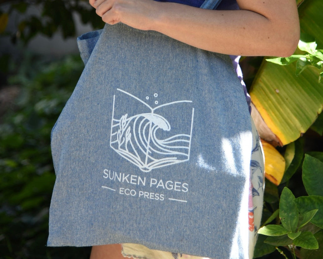 A person outdoors carrying a large heathered blue tote bag printed with a white wave logo and the text 'SUNKEN PAGES ECO PRESS'.